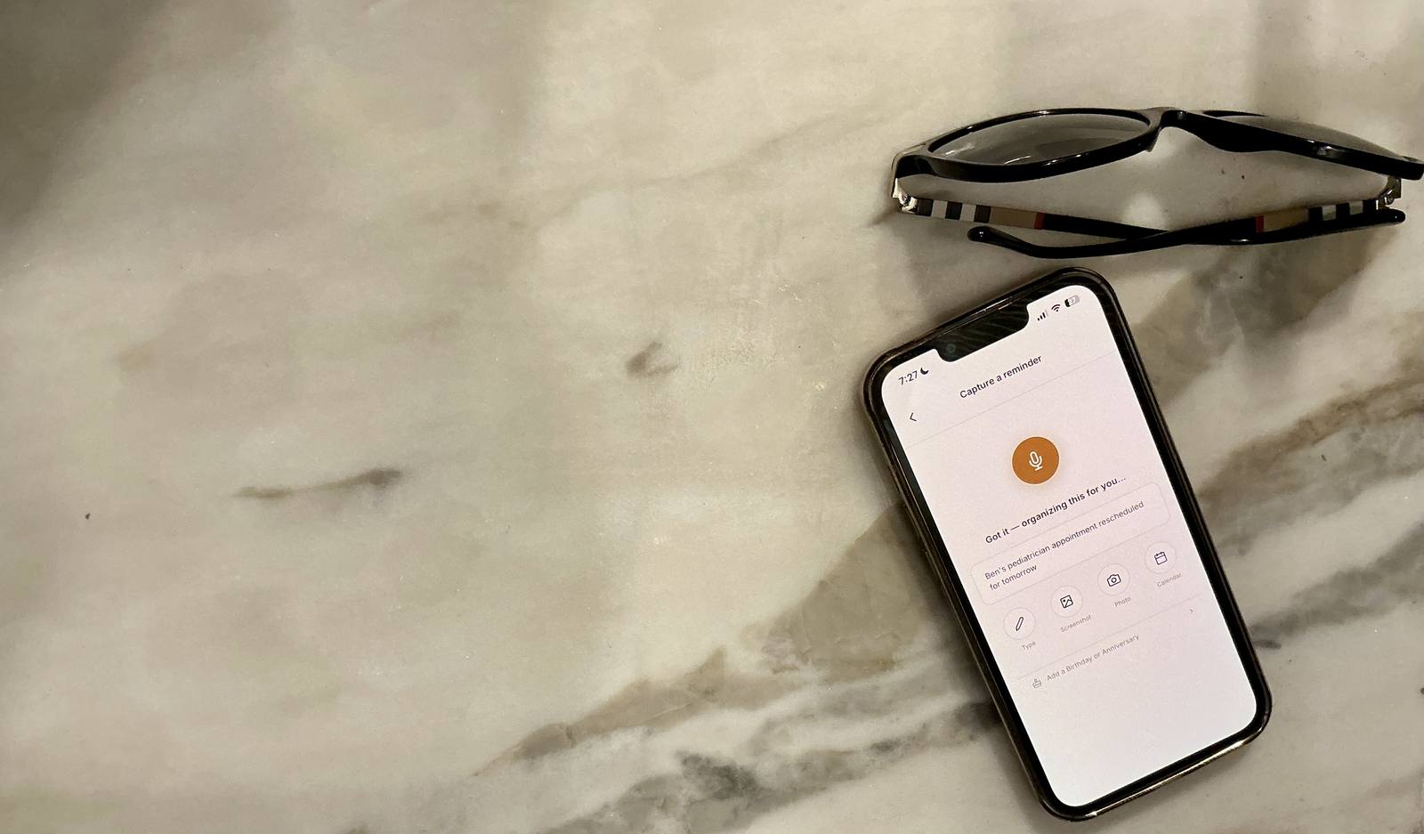 A phone on a marble kitchen counter showing Capture taking a spoken reminder, reading glasses beside it